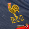 Gros plan sur le coq doré brodé du logo FFF sur le maillot de la France 1998