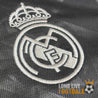 Zoom sur l’écusson brodé du Real Madrid en blanc sur fond gris foncé, avec motif discret en arrière-plan